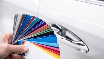 Hand holding vehicle color palette fan with automotive paint samples in white, silver, blue, red, yellow, and black against white car